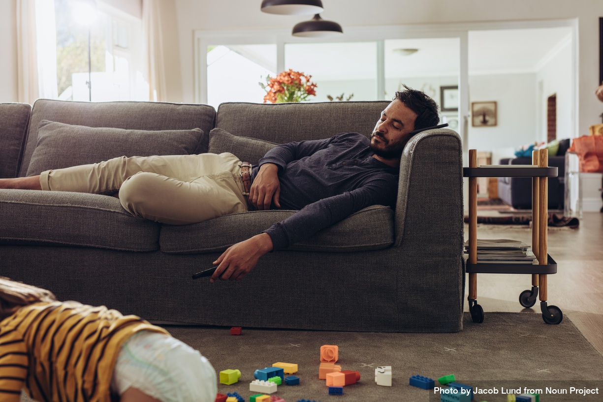 Far i sofaen, mens barn leger på gulvet. Foto: Jacob Lund, Noun Project